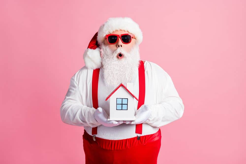 Homme déguisé en pere noel qui porte des lunettes de soleil rouge et tient dans les mains une petite maison.